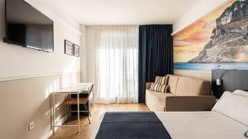 A hotel room with a bed, a desk, a sofa, a TV, and a painting of a coastal scene.