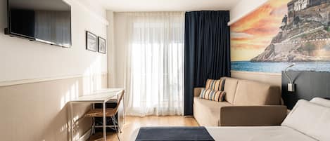 A hotel room with a bed, a desk, a sofa, a TV, and a painting of a coastal scene.