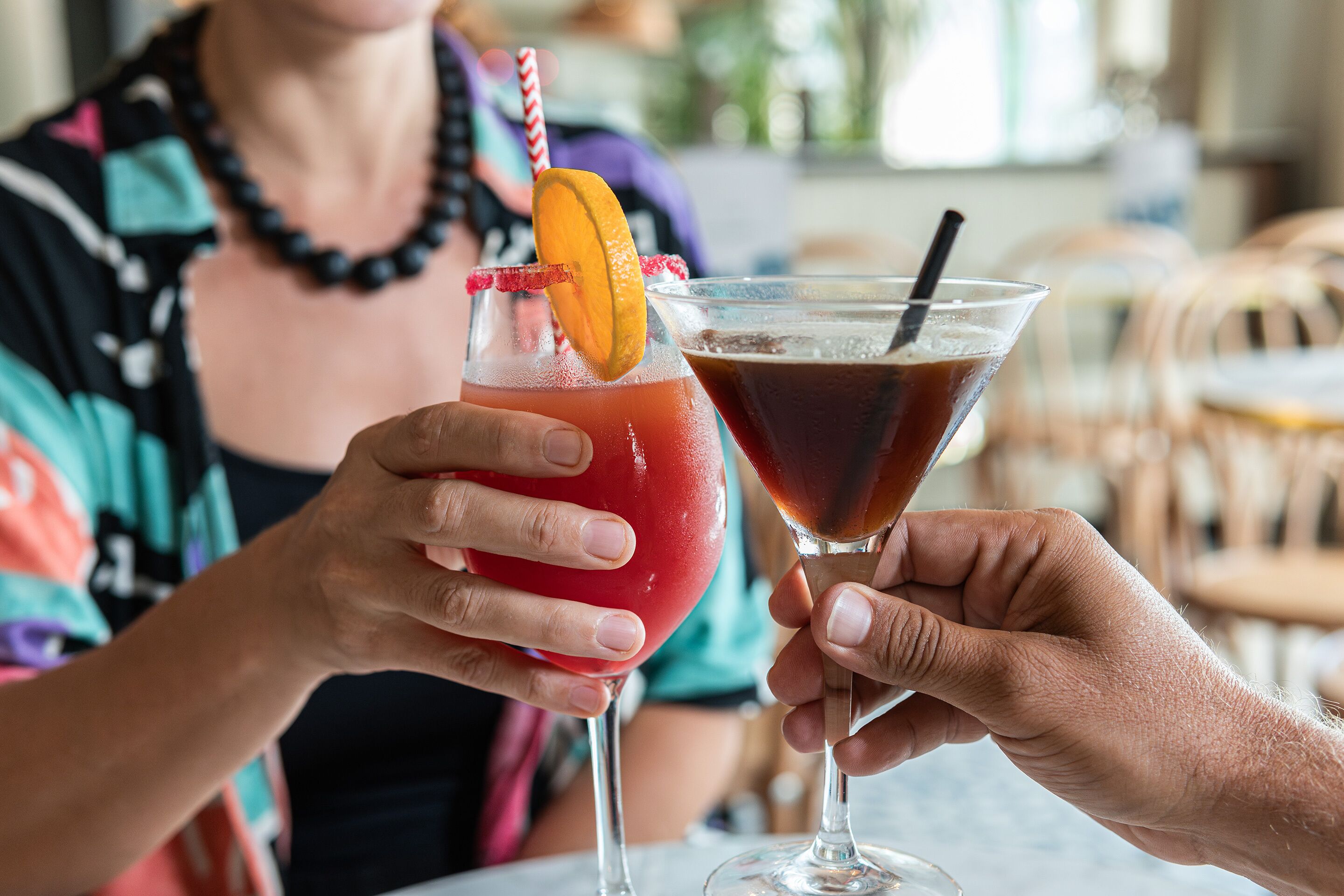 Two people holding cocktail glasses with straws and garnishes.