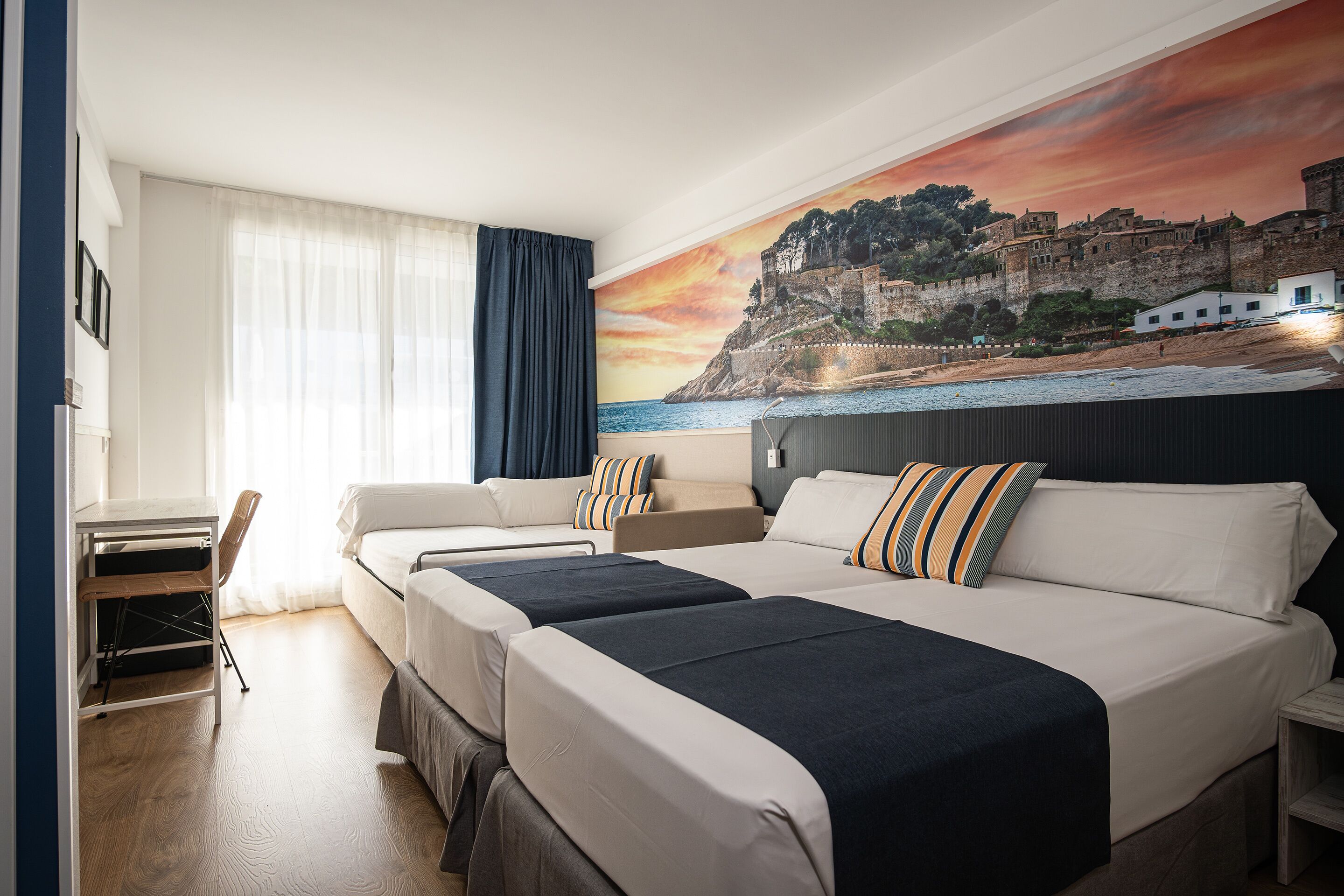 A hotel room with two beds, a desk, and a large scenic mural of a coastal town.