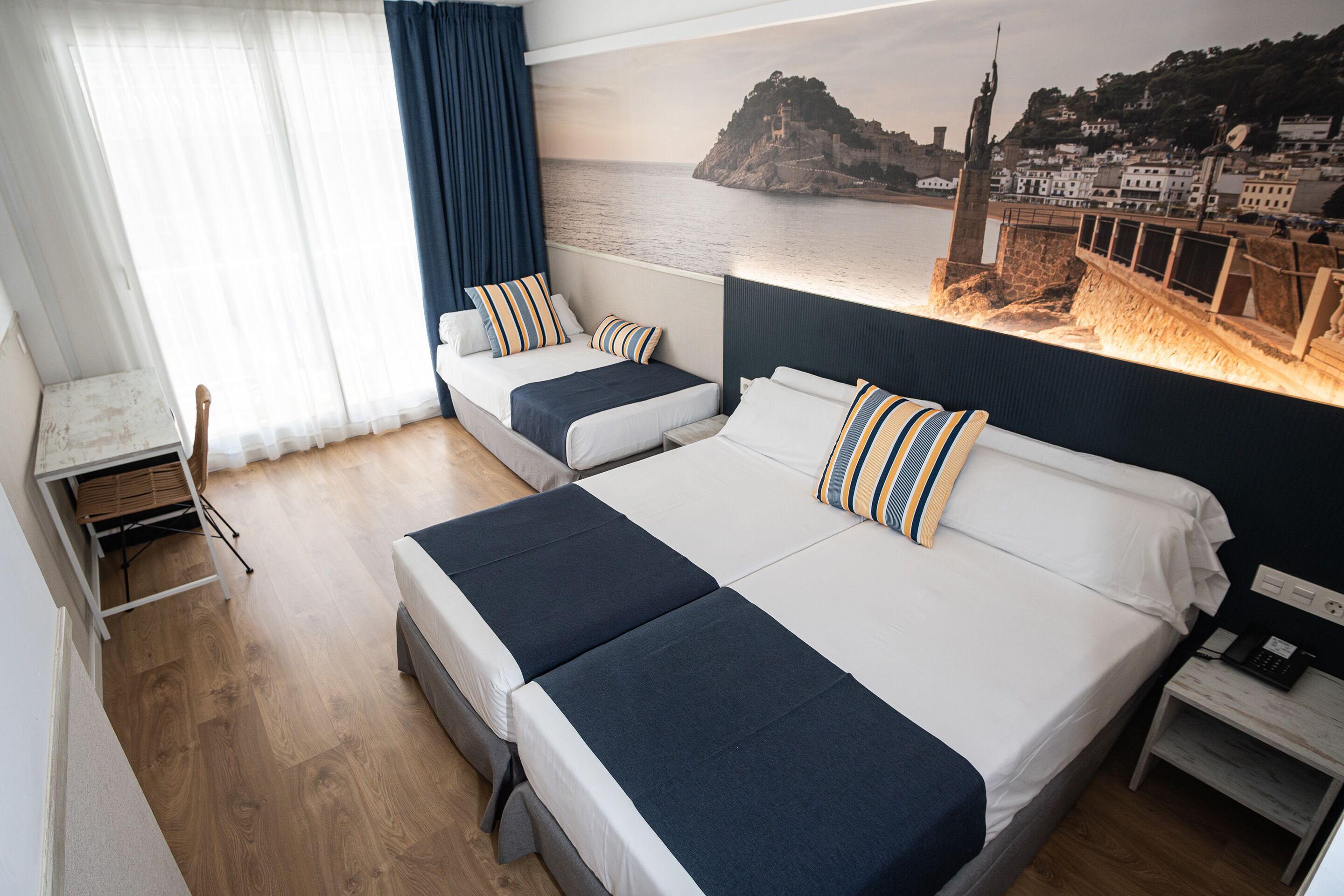 A hotel room with two beds, a desk, and a view of a coastal town.