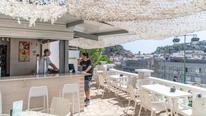 Un café al aire libre con sillas y mesas blancas, una zona de bar y vistas a la ciudad.