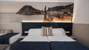 Habitación de hotel con dos camas, vista a un pueblo costero y una estatua en un acantilado.