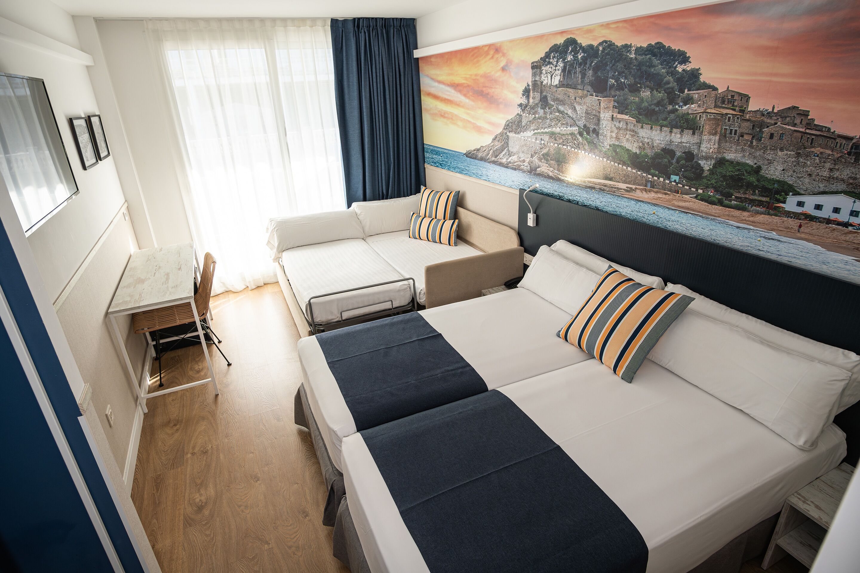 A hotel room with a large bed, a smaller sofa, a desk, and a view of a coastal town.