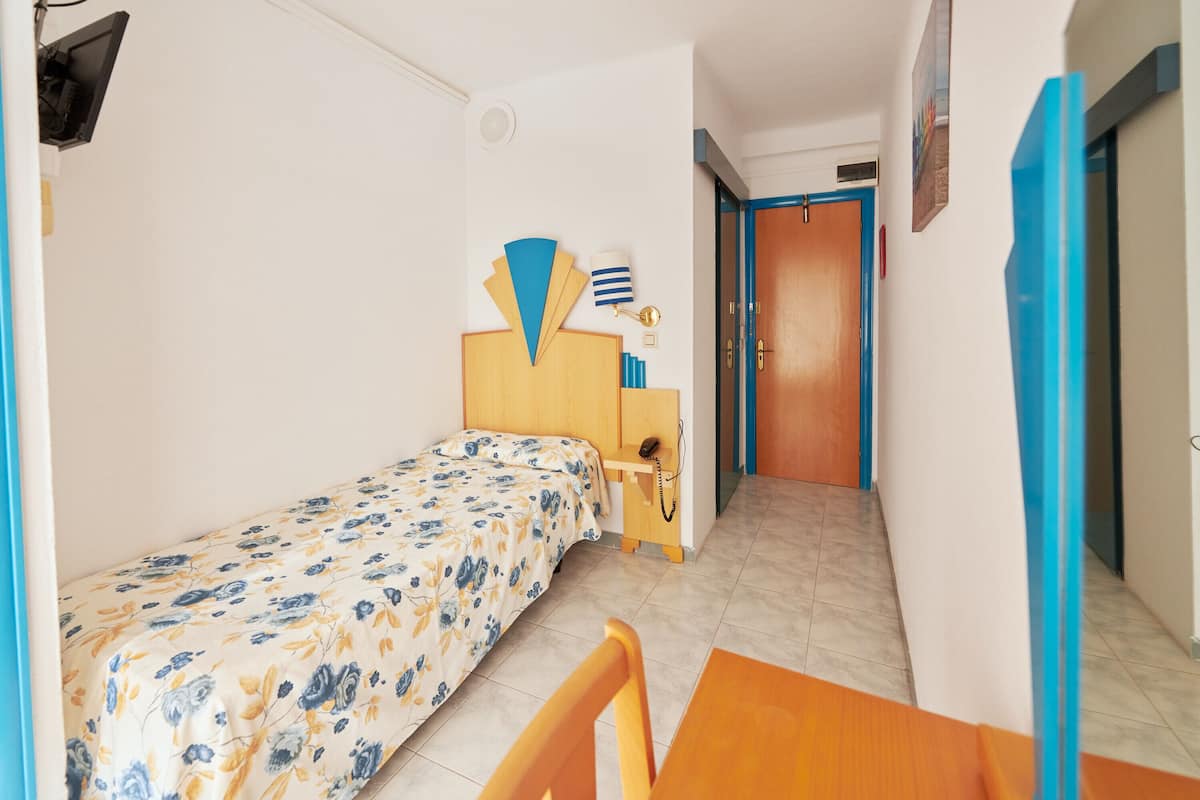 Una habitación pequeña con una cama individual, cabecera de madera, mesita de noche con teléfono y una silla. Una puerta da a un pasillo con marco azul.