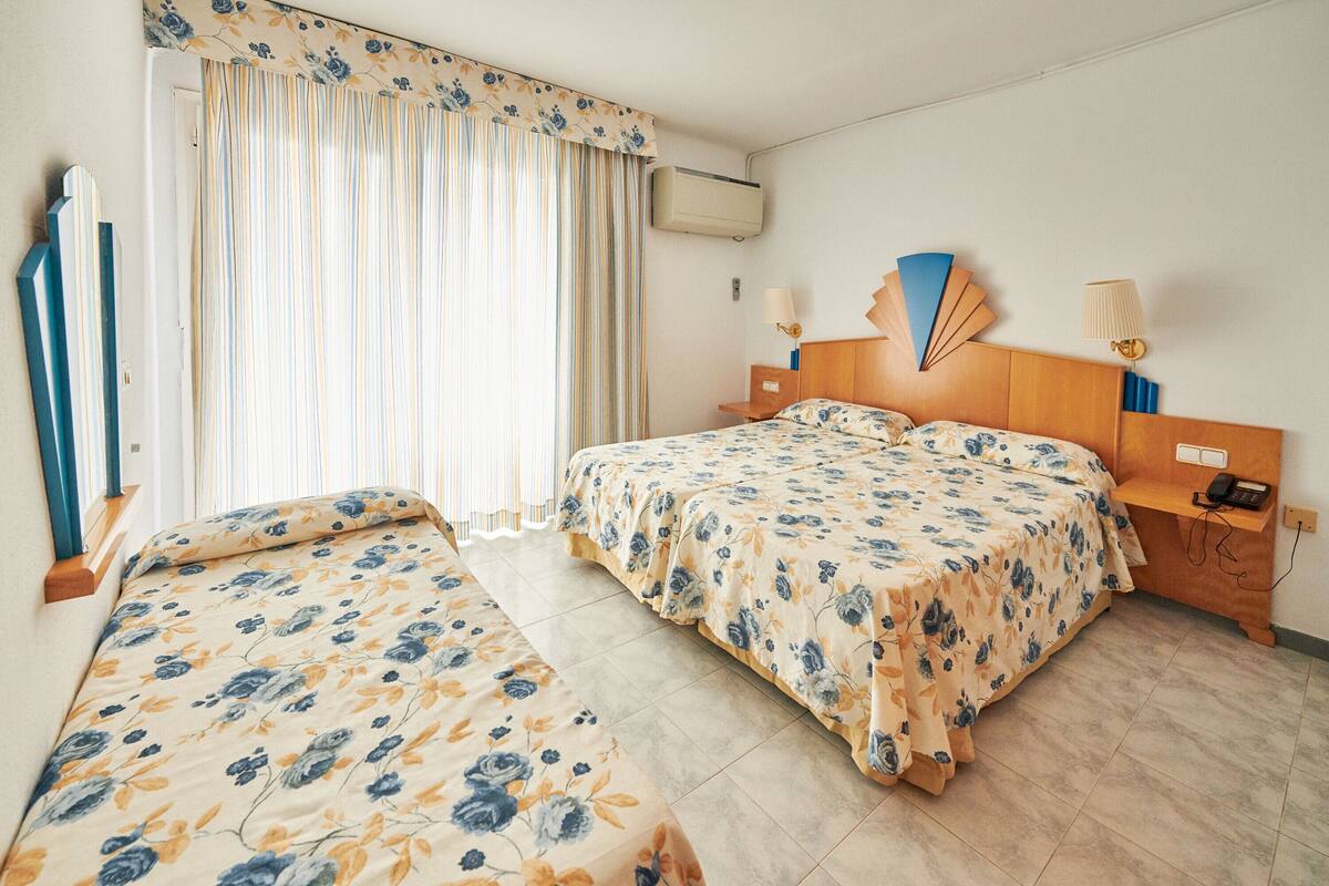 Habitación de hotel con dos camas, cabecera de madera, mesita de noche con lámpara y ventana con cortinas.