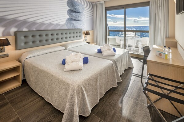 Ein Hotelzimmer mit zwei Betten, einem Schreibtisch, einem Sessel und Blick auf das Meer.