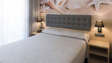 Habitación de hotel con cama, dos mesitas de noche con lámparas, una pared con mural de caracoles y estrellas de mar, y una ventana con cortinas.