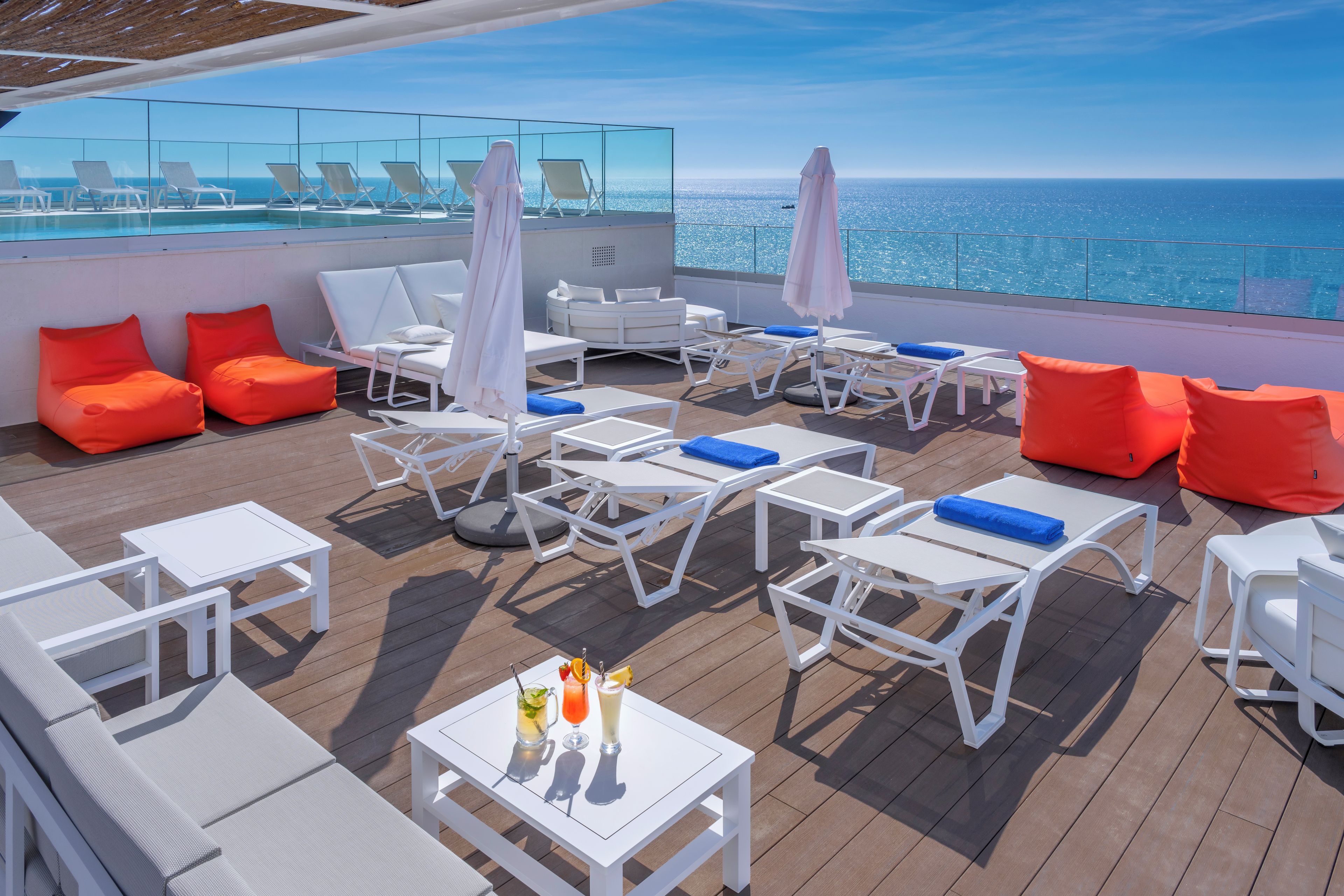 Terraza soleada con sillones blancos, cojines naranjas y una mesa con bebidas.