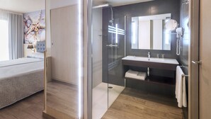 Una habitación de hotel moderna con una cama, ducha con cerramiento de vidrio, baño con lavamanos y espejo, y una obra de arte grande en la pared.