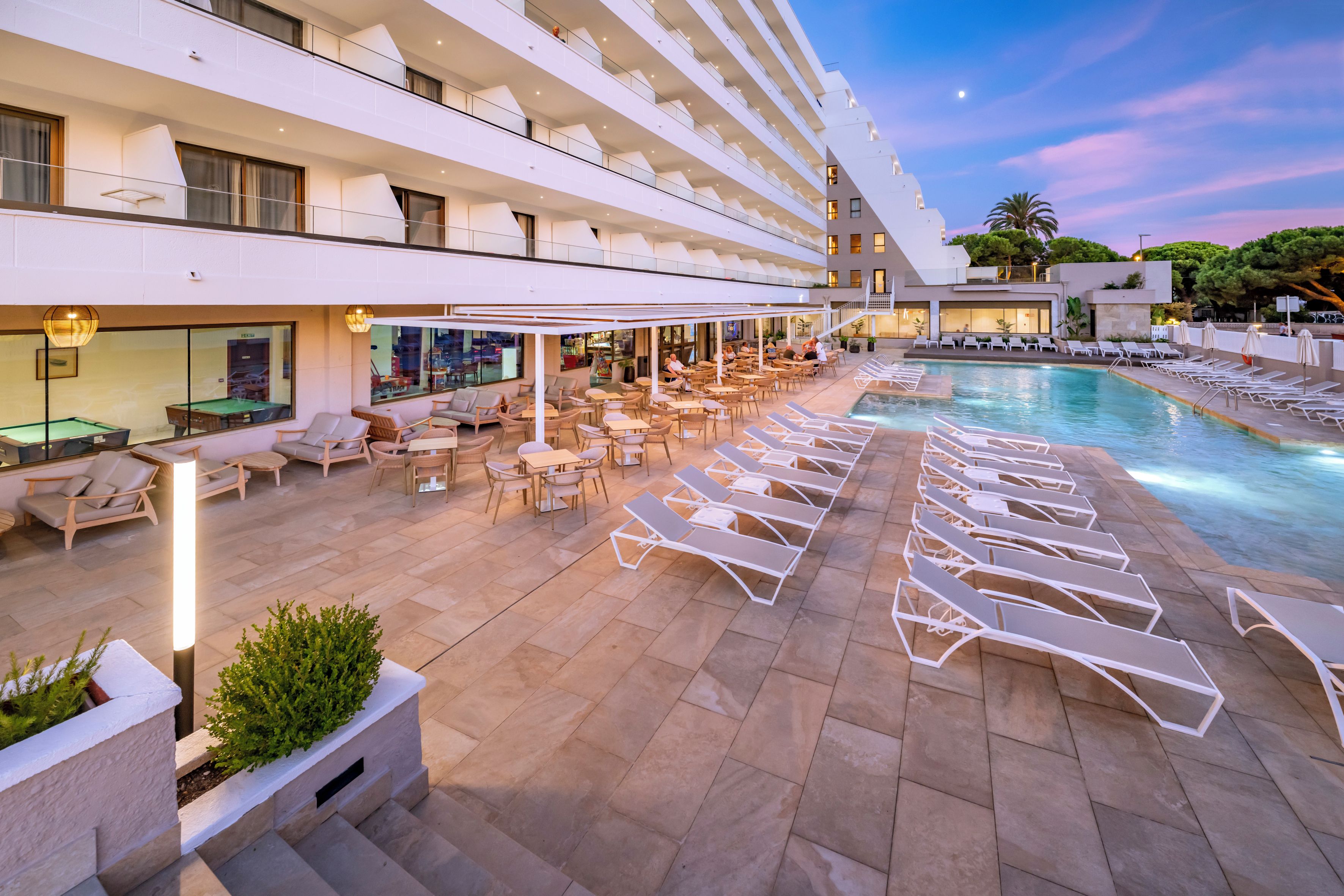Zona de piscina de hotel con sillones de descanso, mesas y una piscina.