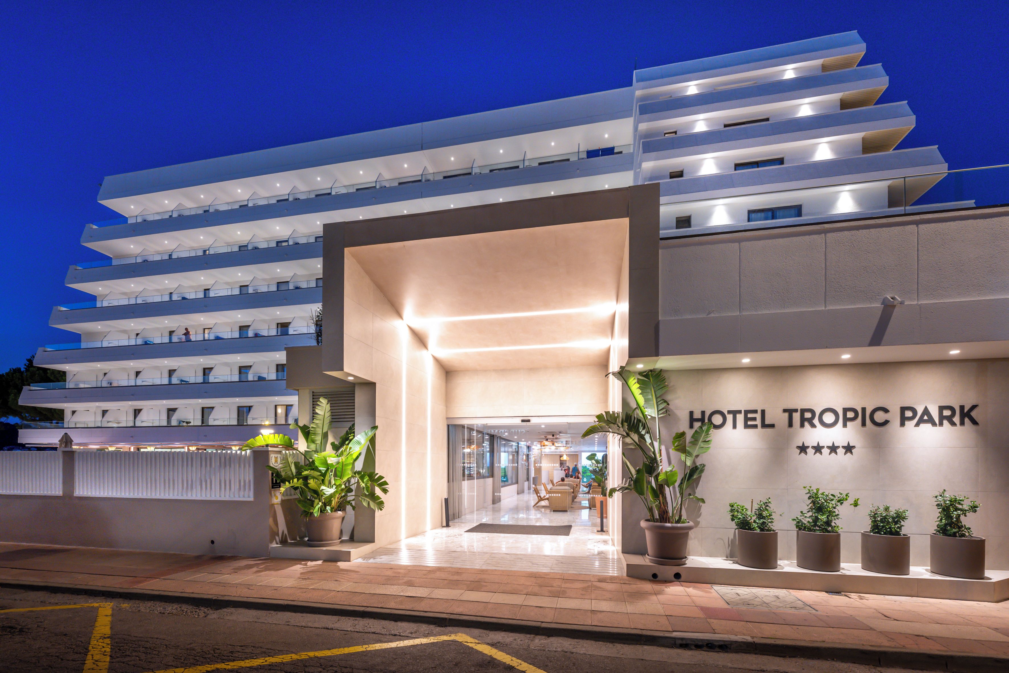 A modern hotel entrance with a clear sign reading 'HOTEL TROPIC PARK' and a series of potted plants at the entrance.