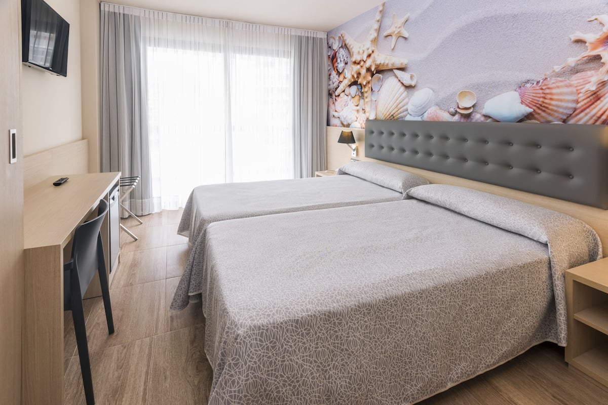 Ett hotellrum med två sängar, ett skrivbord, en stol och en stor väggmålning med motiv av snäckor och sjöstjärnor.