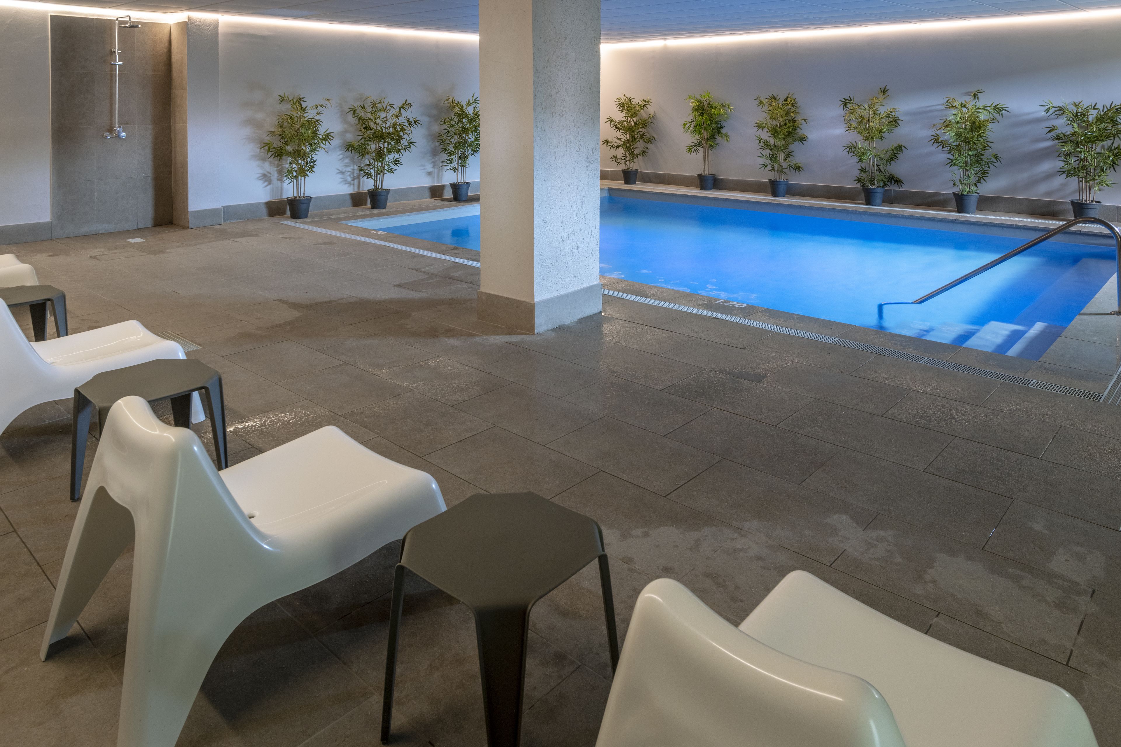 Uma área de piscina com piso ladrilhado, várias cadeiras brancas e pretas e plantas em vasos ao longo da borda da piscina.