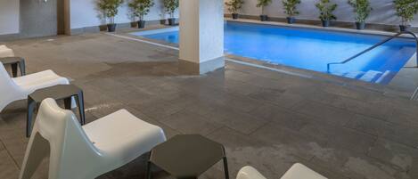 A swimming pool area with a tiled floor, several white and black chairs, and potted plants along the poolside.