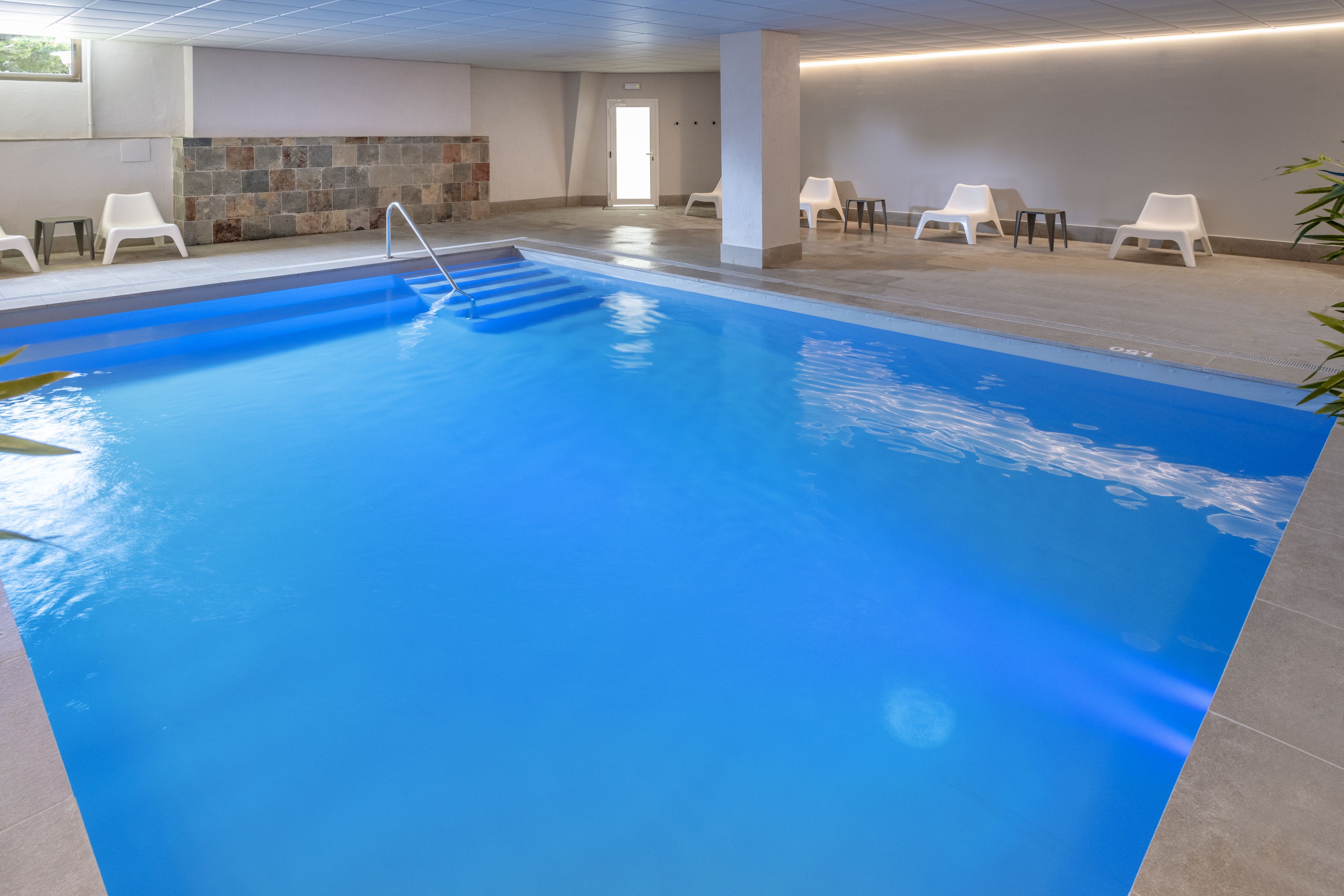 An indoor swimming pool with clear water, a diving board, and several white lounge chairs around the pool area.