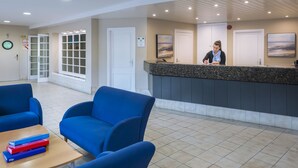 A reception area with a receptionist, blue chairs, a reception desk, and a view of the outside. - Apart-hotel GHT Tossa Park (Tossa de Mar)