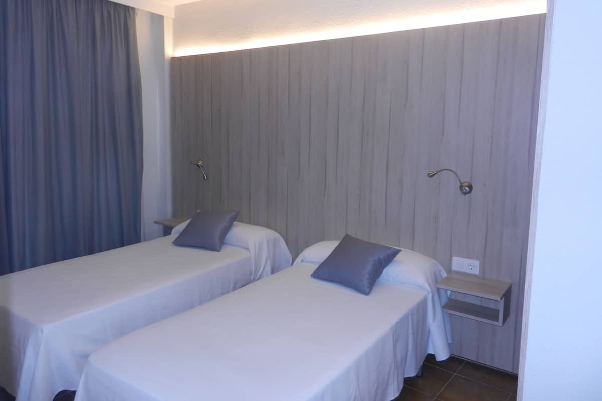 Quarto de hotel com duas camas de solteiro, uma mesinha de cabeceira, uma luminária na parede e uma cortina.