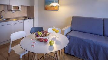 Una cocina compacta con microondas, pileta y alacenas. Una mesa de comedor con sillas y un bol de frutas está en el centro. Un sofá azul está apoyado contra la pared.