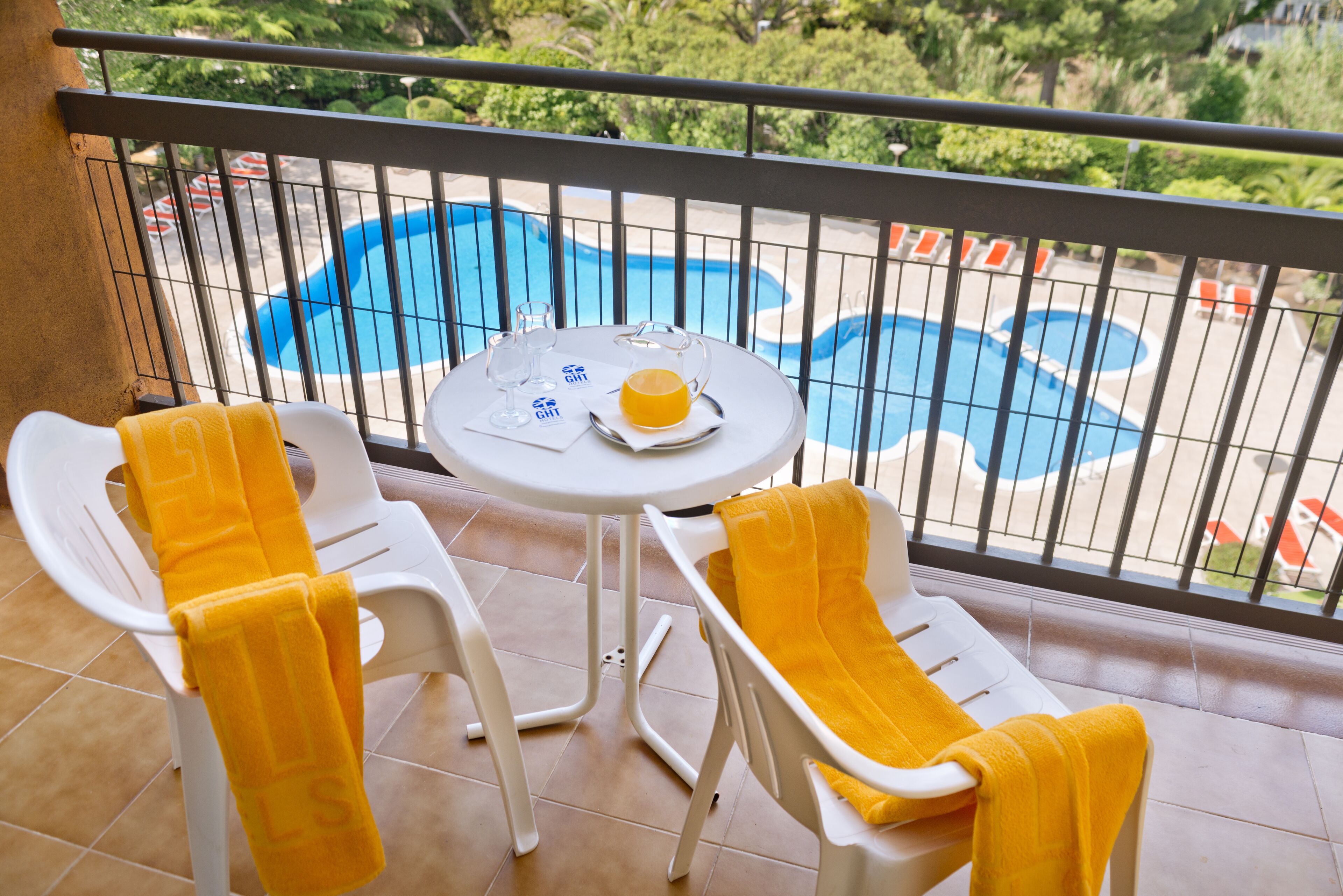 Un balcon donnant sur une piscine, une table avec un verre de jus d’orange et deux chaises avec des serviettes jaunes.