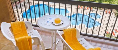 Een balkon met uitzicht op een zwembad, een tafel met een glas sinaasappelsap en twee stoelen met gele handdoeken.