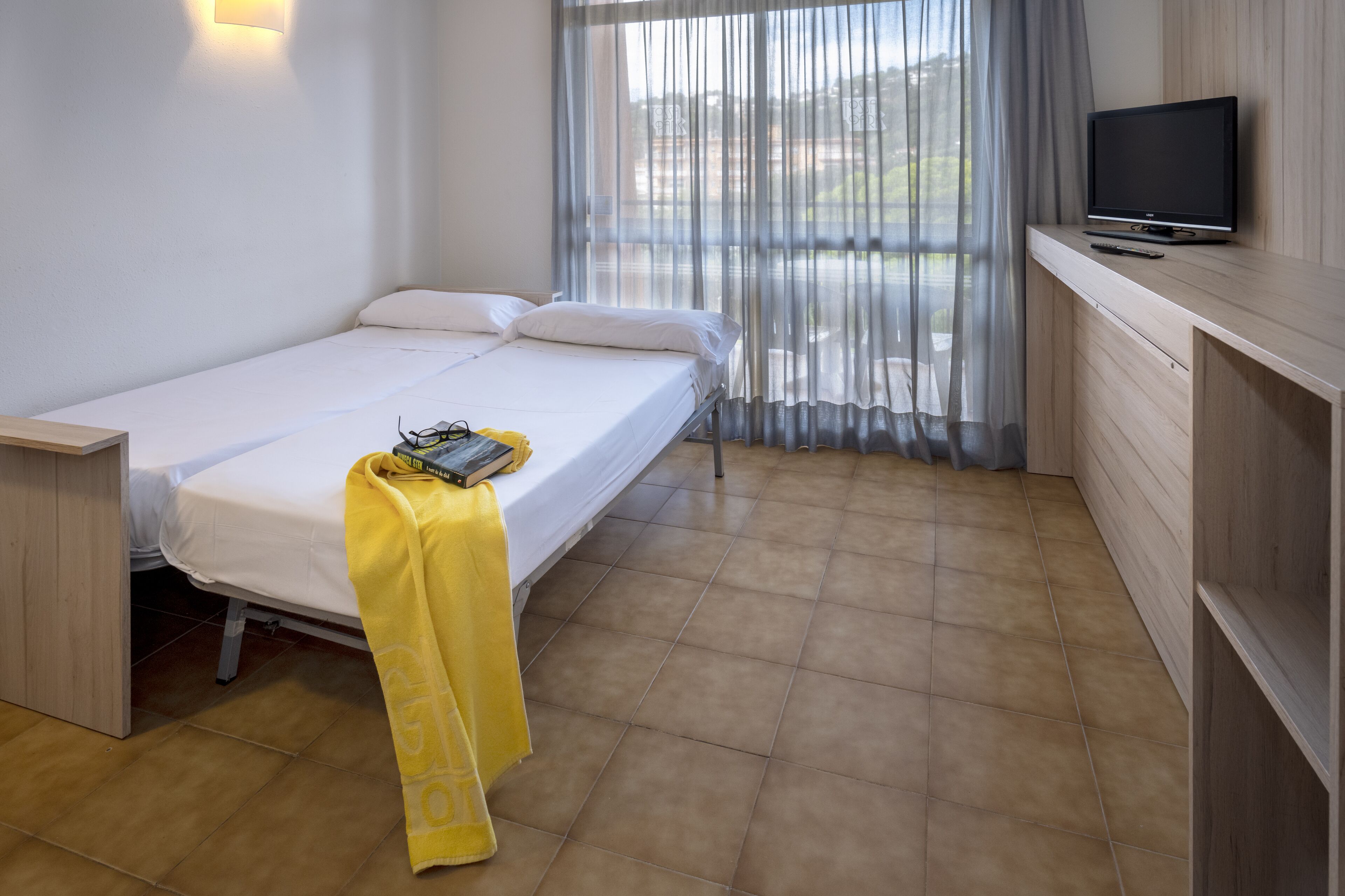 A single-bed room with a TV on a wooden stand, a bedside table, and a window with sheer curtains.