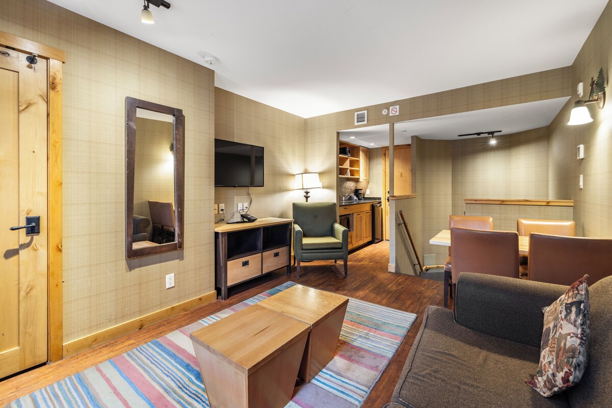A modern living room with a flat-screen TV, a sofa, a coffee table, and a small kitchen area.