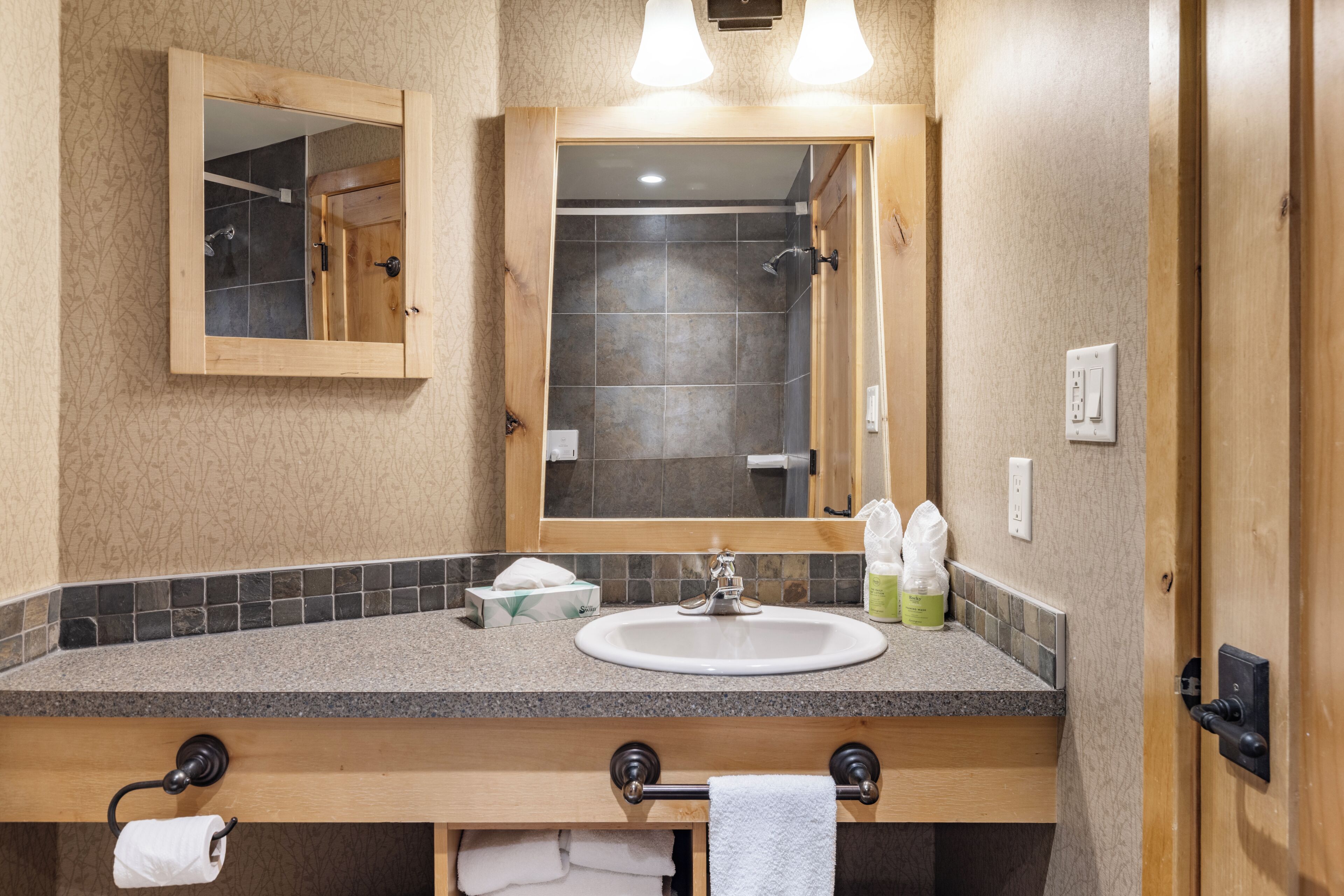 A bathroom with a wooden vanity, a sink, a mirror, and a shower visible through the door.
