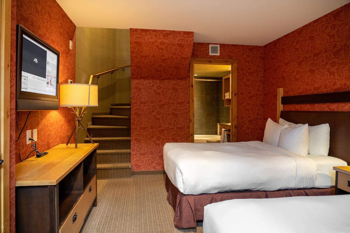A hotel room with two beds, a desk with a lamp, a TV, and a staircase leading to a bathroom.