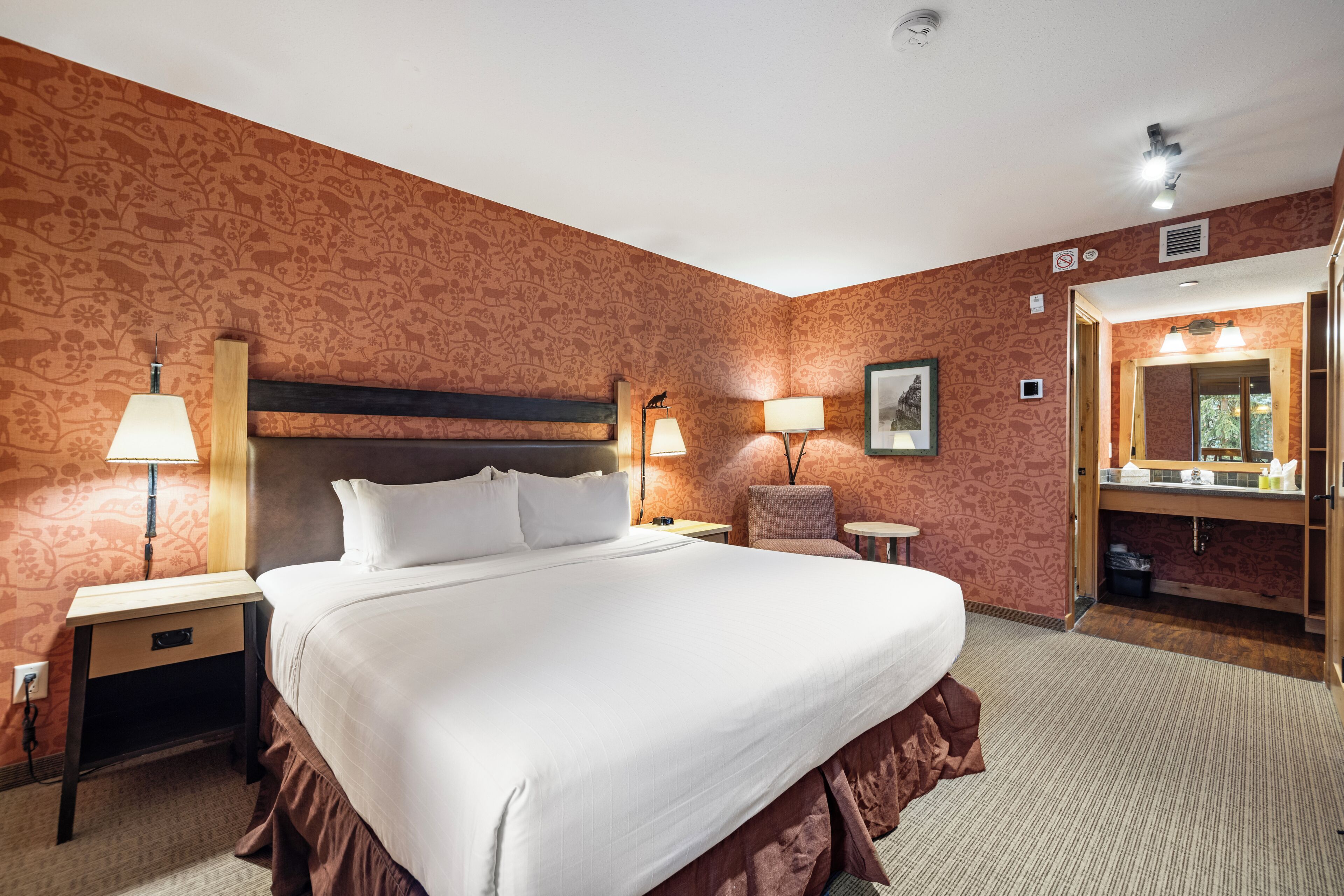 A hotel room with a large bed, two bedside tables, a chair, a mirror, and a bathroom visible through an open door.