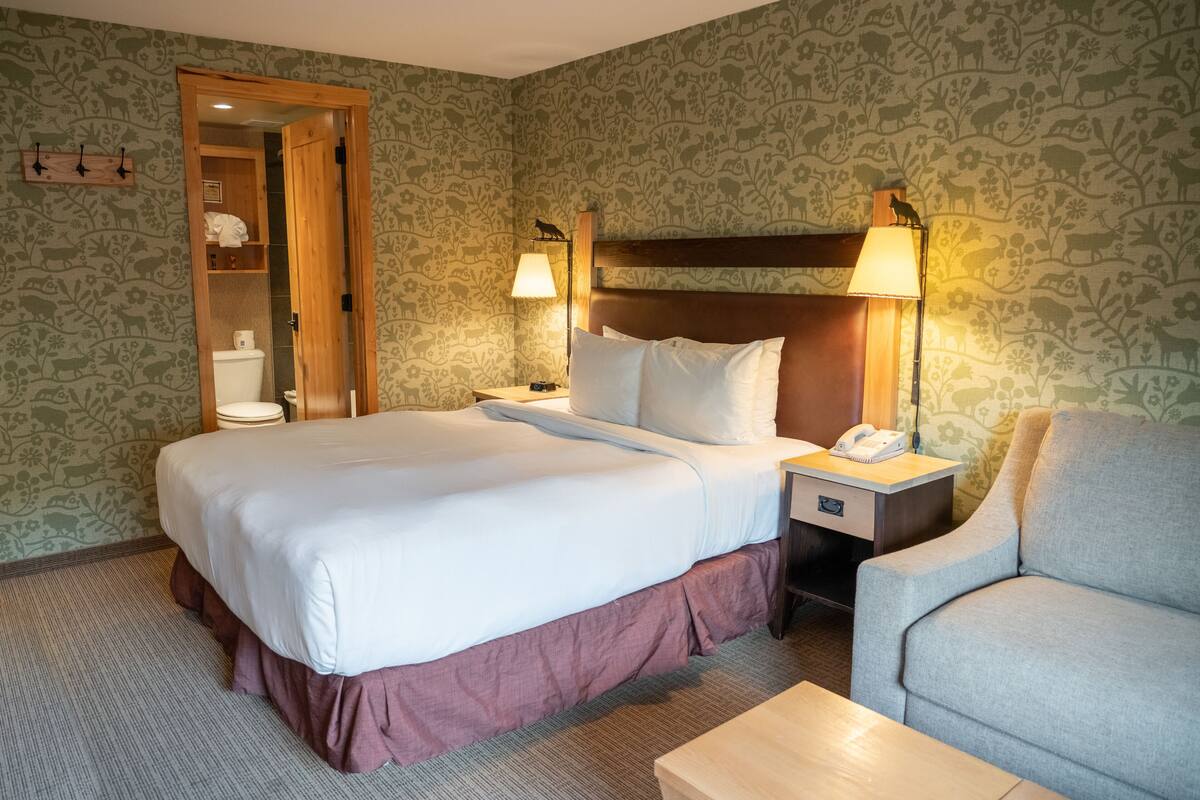A hotel room with a large bed, a wooden headboard, two bedside lamps, a desk with a telephone, a grey armchair, and a wooden cabinet with toiletries.