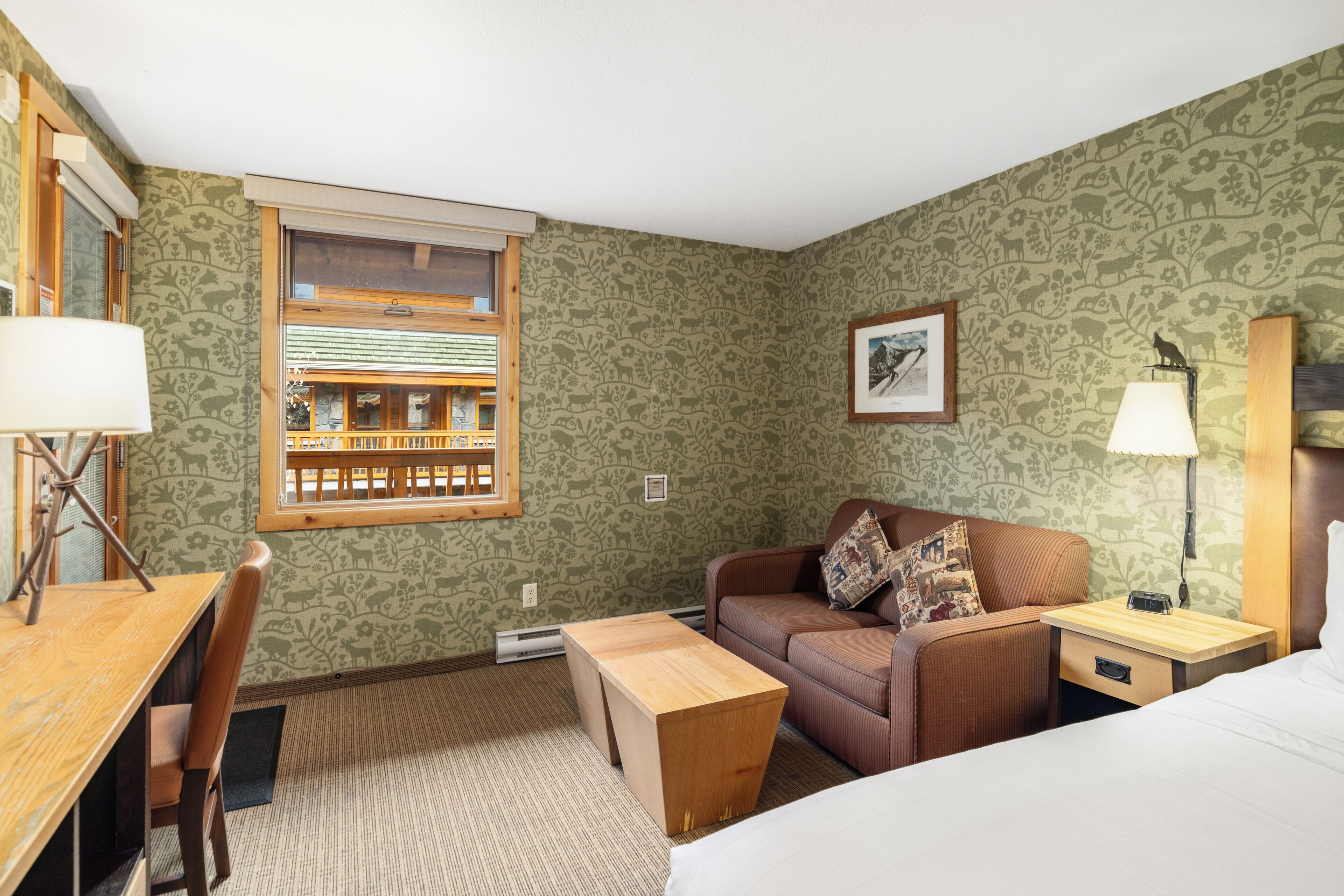 A room with a wooden desk, a brown sofa, a bed, and a window with a view of a balcony.