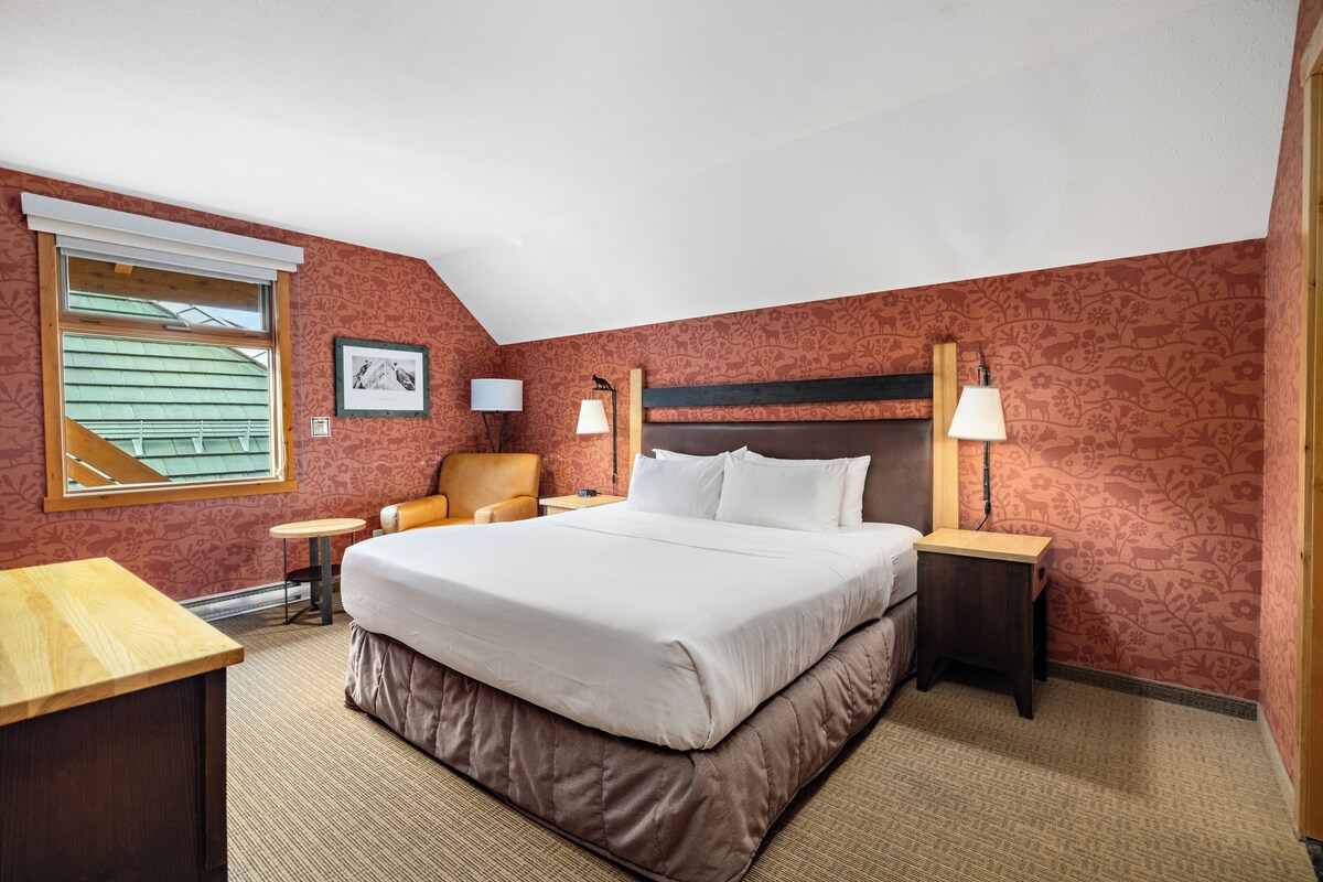 A hotel room with a large bed, a chair, a table, and a lamp.