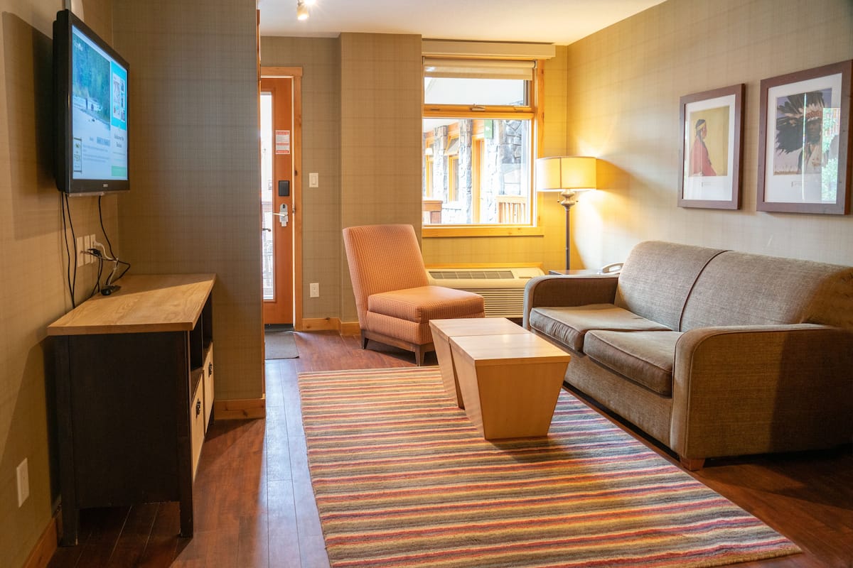 A living room with a sofa, armchair, and coffee table, a TV mounted on the wall, and a window with a view of buildings.