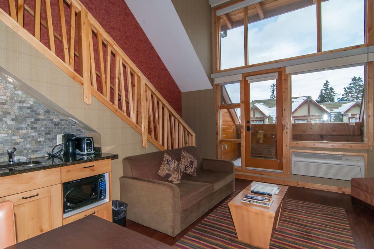 A cozy living area with a wooden staircase, a kitchen with a microwave, and large windows offering a view of houses and trees.