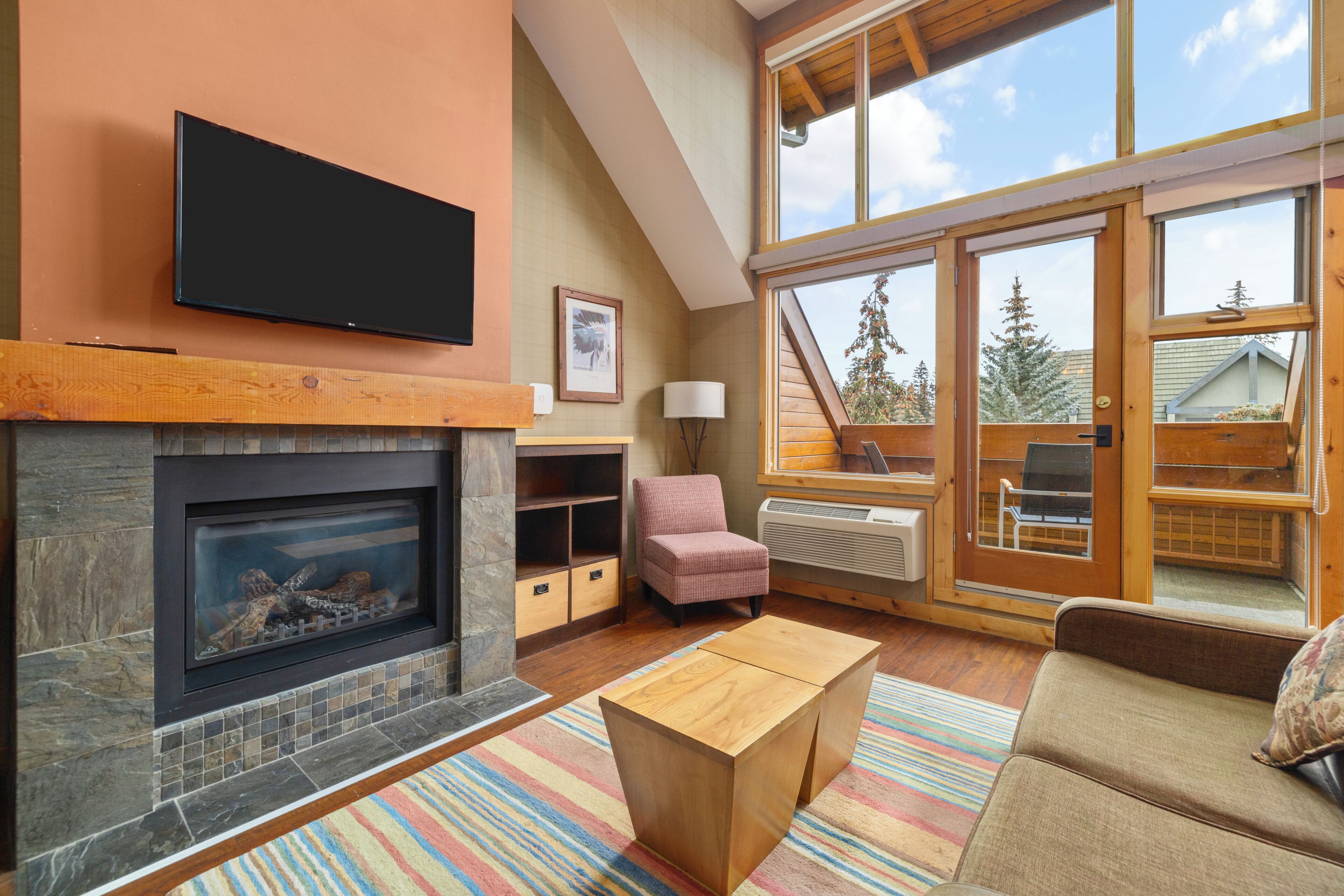 A living room with a fireplace, a television mounted above it, a sofa, a chair, and a window with a view of trees.
