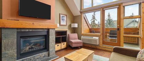 A living room with a fireplace, a television mounted above it, a sofa, a chair, and a window with a view of trees.