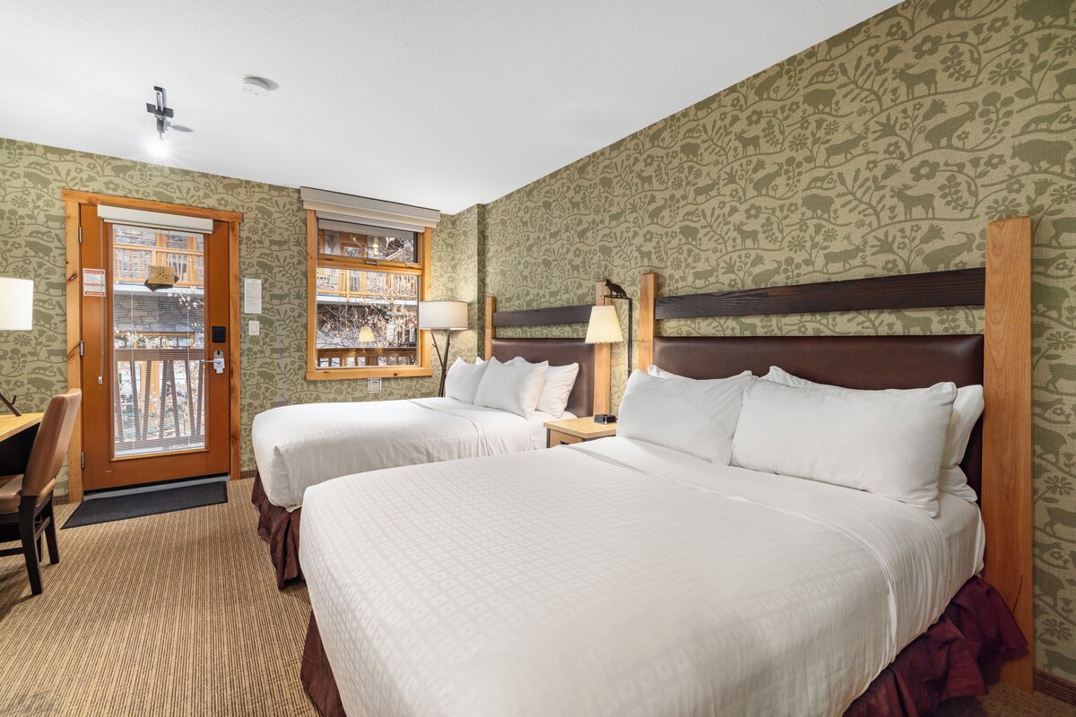 A hotel room with two beds, a desk, a chair, a window with blinds, and a door.