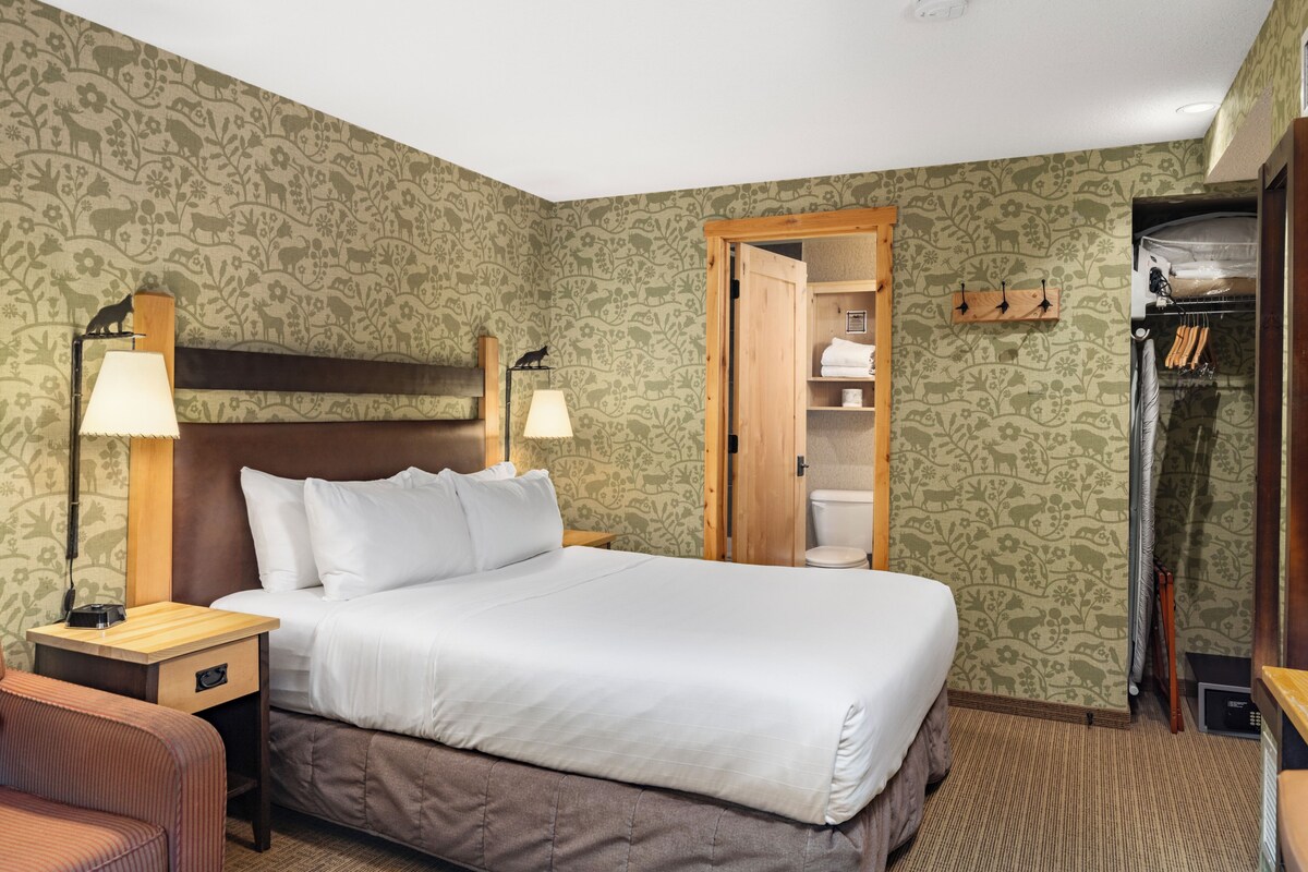 A hotel room with a bed, bedside lamps, a wooden headboard, a wardrobe, and a bathroom with a toilet and shower.