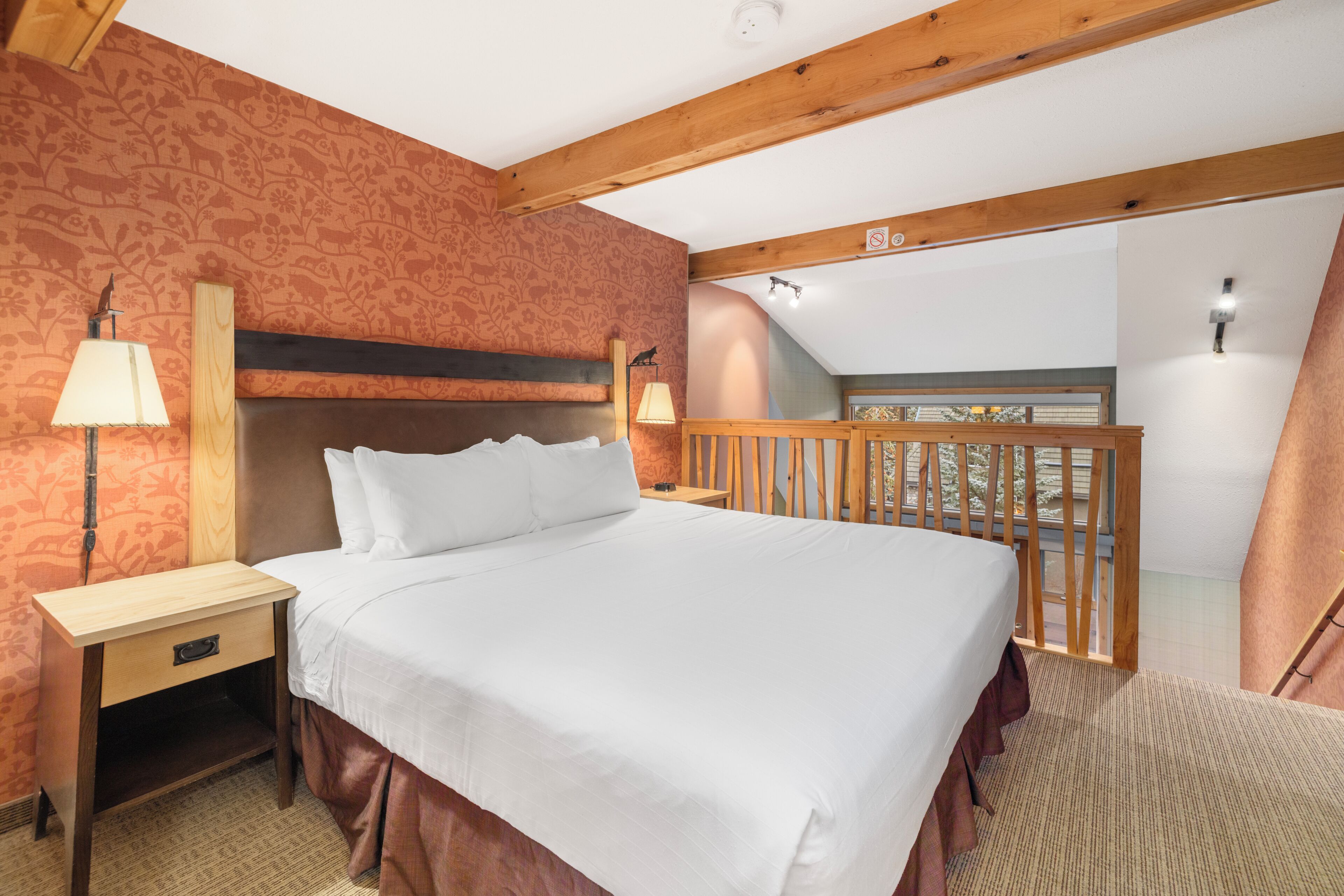 Habitación de hotel con cama, mesitas de noche, lámpara, cabecero de madera, balcón con barandilla y pared con diseño.
