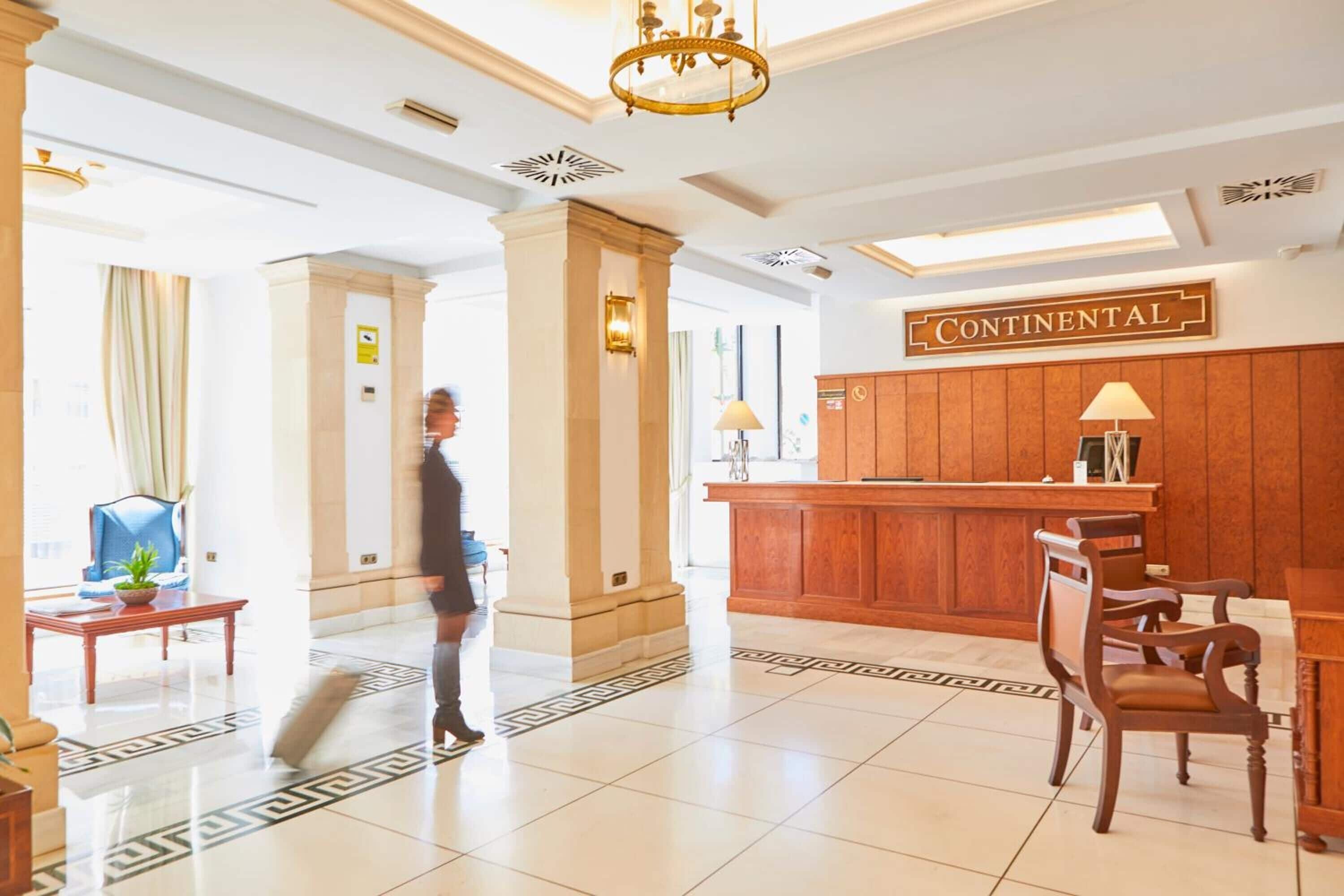 Un hall d’hôtel avec un comptoir d’accueil, des chaises en bois, un lustre et une enseigne indiquant  « Continental ».