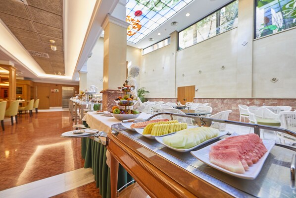 Zona de bufé con diversas opciones de comida, incluyendo frutas y verduras, en un espacio interior bien iluminado con piso de mármol y decoración moderna.