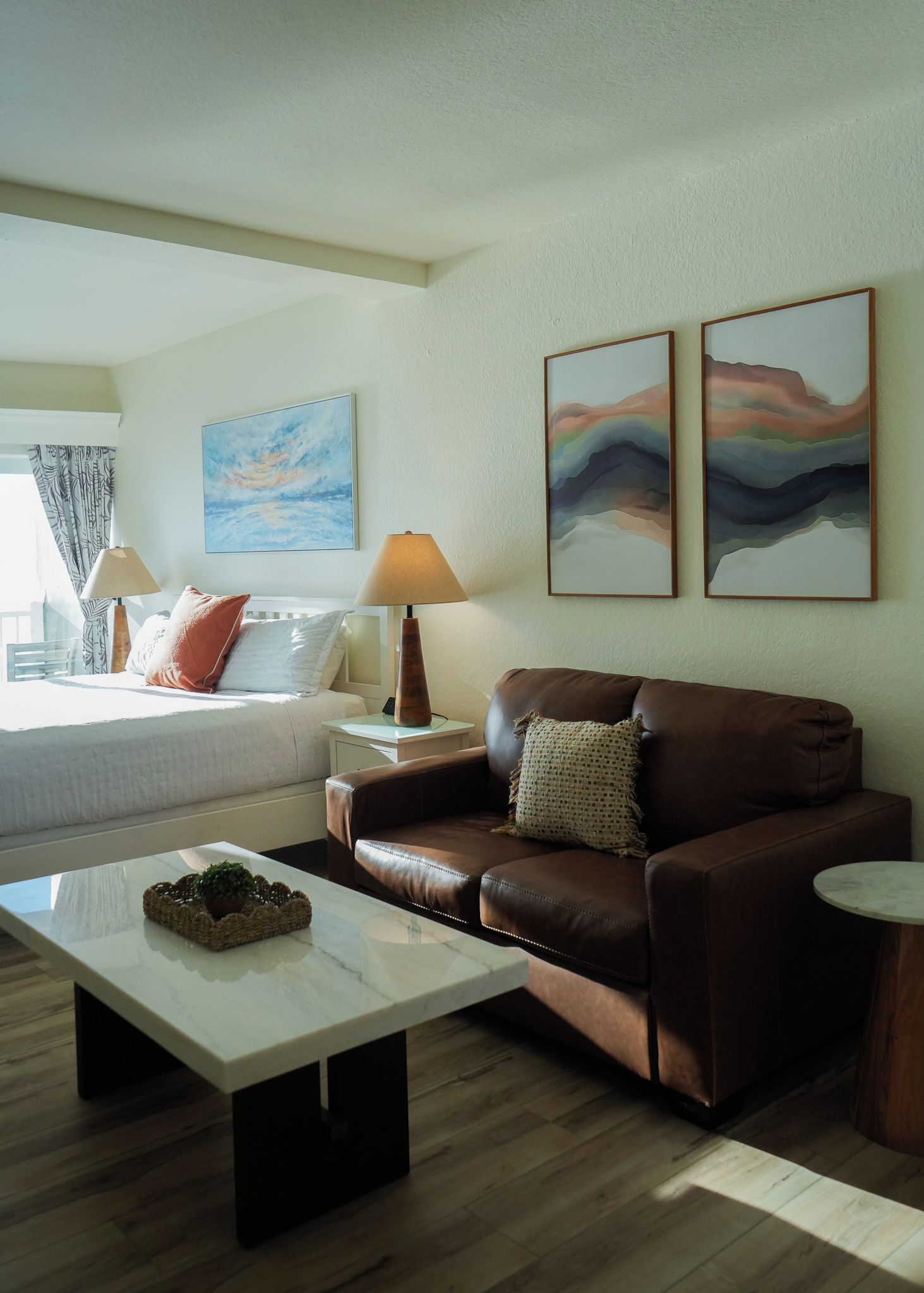 Una habitación de hotel con una cama, un sofá de cuero marrón, una mesita y dos cuadros abstractos en la pared.