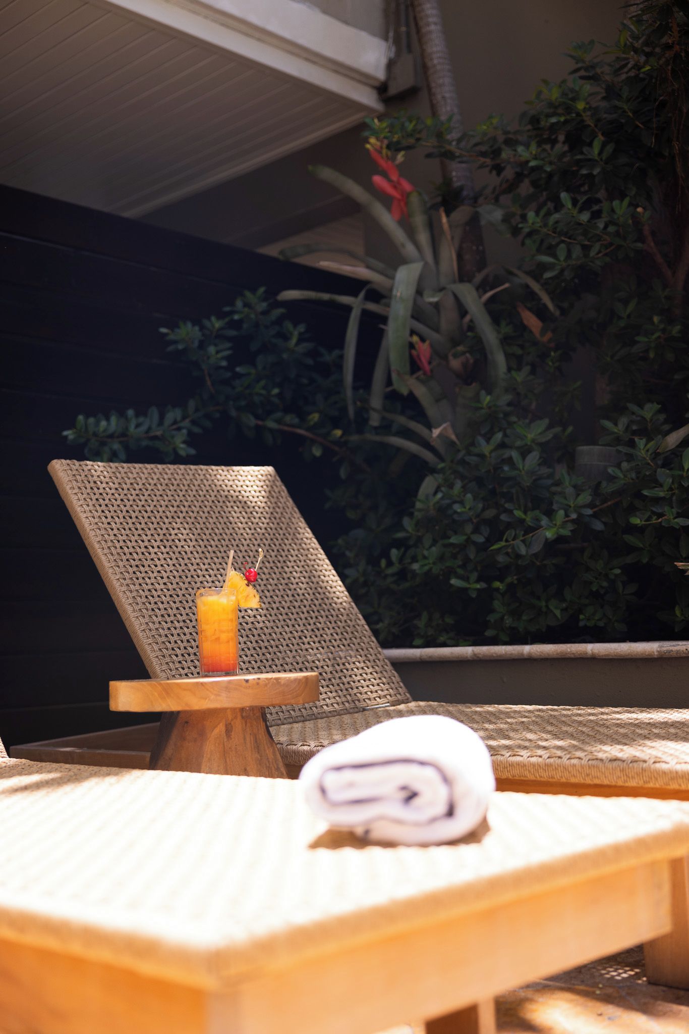 A lounge chair with a towel, a glass of orange juice, and a cocktail with a cherry on a small wooden table.