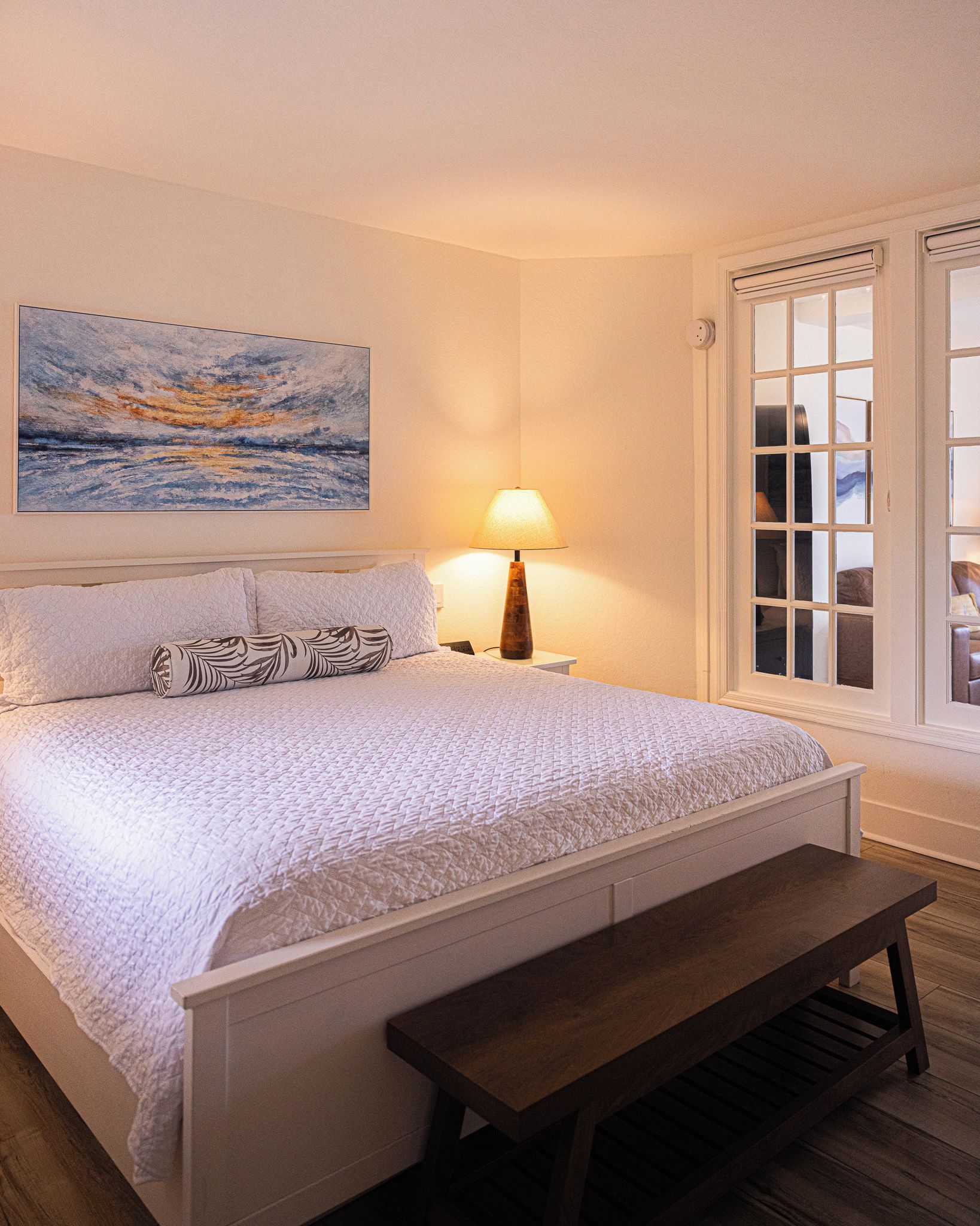 Ein Schlafzimmer mit einem Bett, einer Bank, einem Nachttisch mit Lampe, einem Fenster mit Jalousien und einem Gemälde an der Wand.
