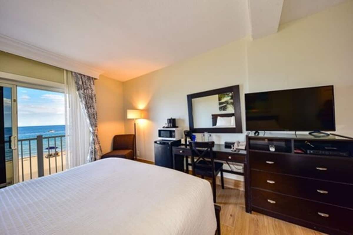 A hotel room with a bed, a desk, a dresser, a TV, a lamp, and a view of the sea.