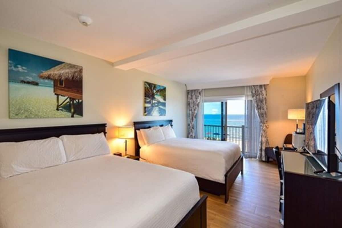 A hotel room with two beds, a desk, a TV, and a balcony with a view of the sea.