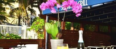 A table with a white mug, a glass of orange juice, and a bottle of ZONIN champagne, set outdoors with a blue awning labeled "GRILL" and pink orchids in the background.