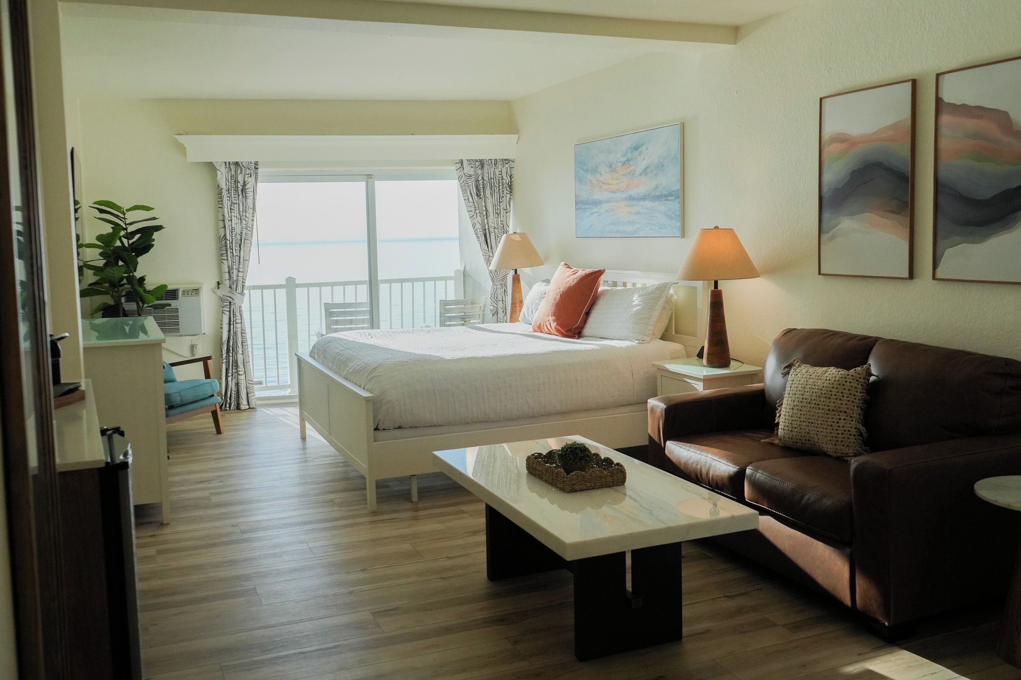 Habitación de hotel con cama, sofá de cuero marrón, una mesita y un balcón con cortinas.