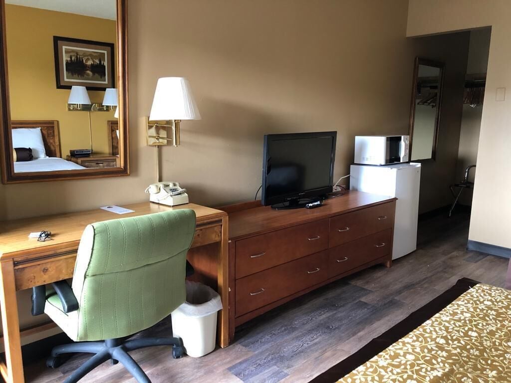 A hotel room with a wooden desk, a green office chair, a television, a dresser, a mirror, and a small refrigerator.